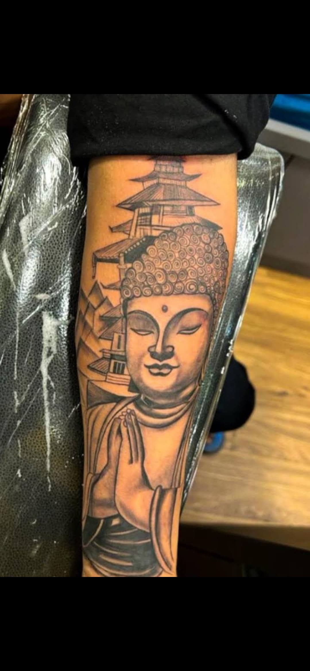 Black and grey forearm tattoo of a praying Buddha in front of a multi-tiered pagoda.