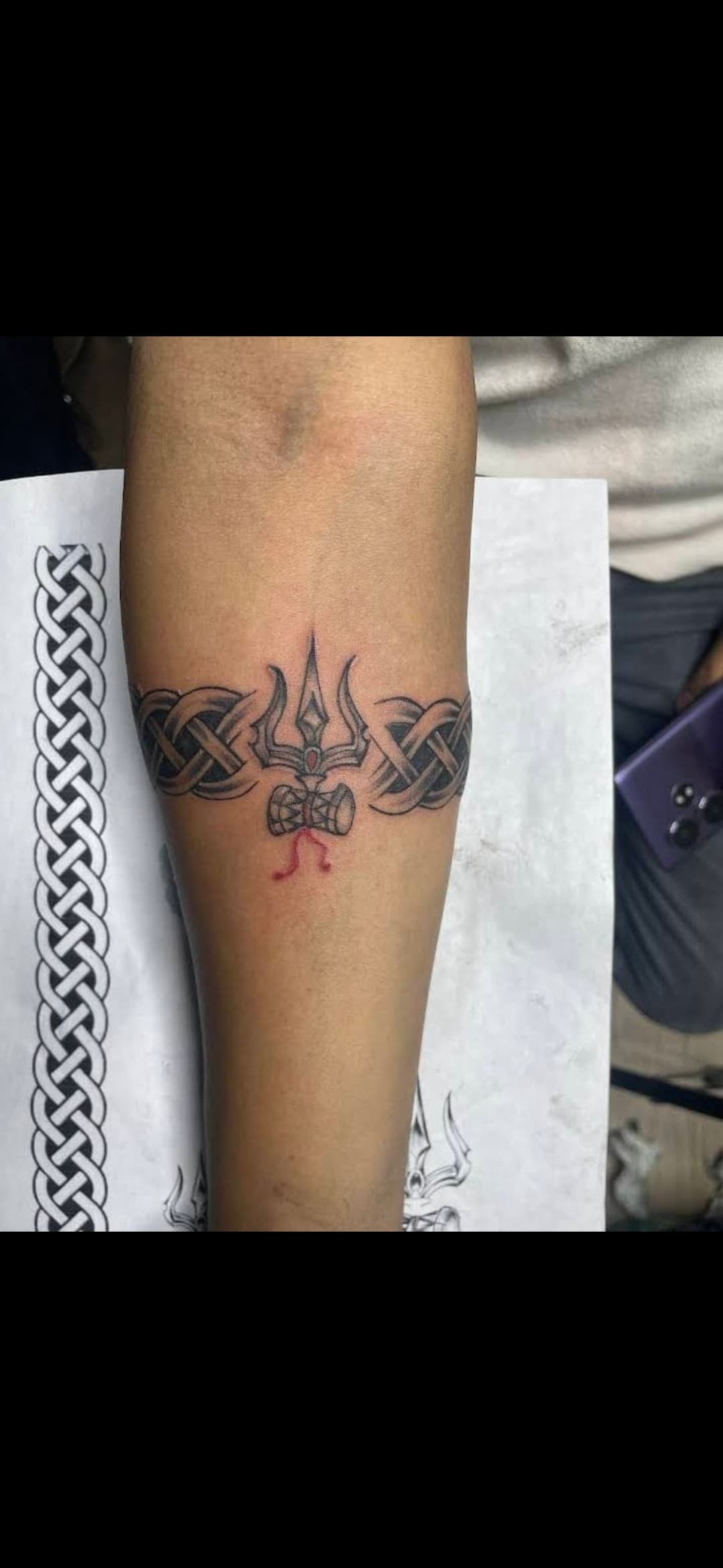 Forearm tattoo featuring a central trishula and damru integrated into a black Celtic knot armband.